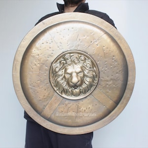 Puede incluir: Un escudo grande y redondo de color bronce con un emblema detallado de cabeza de león en el centro. El escudo tiene una apariencia desgastada con arañazos visibles y una superficie texturizada. El texto "Armorcollectionstore" es visible.
