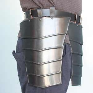Puede incluir: Un par de placas de armadura de pierna de metal, una en cada pierna, usadas sobre un cinturón marrón. Las placas son de metal y tienen un diseño en capas. Las placas se sujetan al cinturón con correas.