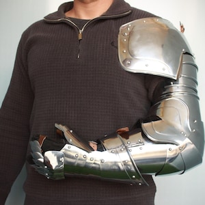 Könnte beinhalten: Eine silberne, artikulierte Armrüstung im mittelalterlichen Stil. Die Rüstung bedeckt den gesamten Arm von der Schulter bis zur Hand, inklusive Handschuh. Die Rüstung besteht aus Metallplatten, die mit Nieten verbunden sind.