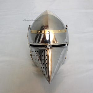 May include: A silver helmet with a pointed visor and a brass band around the top. The helmet has a small, pointed nose guard and a series of small holes in the visor.