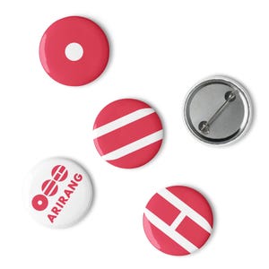 May include: A collection of five round pinback buttons. The buttons feature various designs, including a red button with a white dot, a button with white stripes on a red background, and a button with the text "ARIRANG".