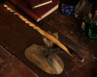 May include: A wooden wand with a twisted design, resting on a piece of driftwood. The wand is light brown with a darker brown swirl pattern.