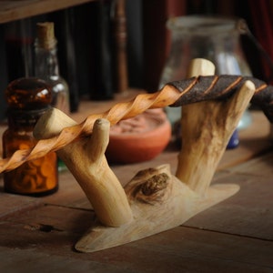 May include: A wooden wand with a dark brown leather wrap is displayed on a wooden stand. The wand is twisted and has a natural wood finish.