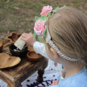 May include: A young person wearing a floral headband with pink roses and green leaves. The person is reaching towards a small wooden table with various wooden bowls and a heart-shaped dish. The table is on a grassy surface.