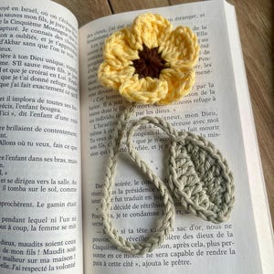May include: A handmade crochet bookmark featuring a yellow flower with a dark brown center, attached to a long, braided light green stem with a single textured leaf. The item is placed on an open book with printed French text on the pages.