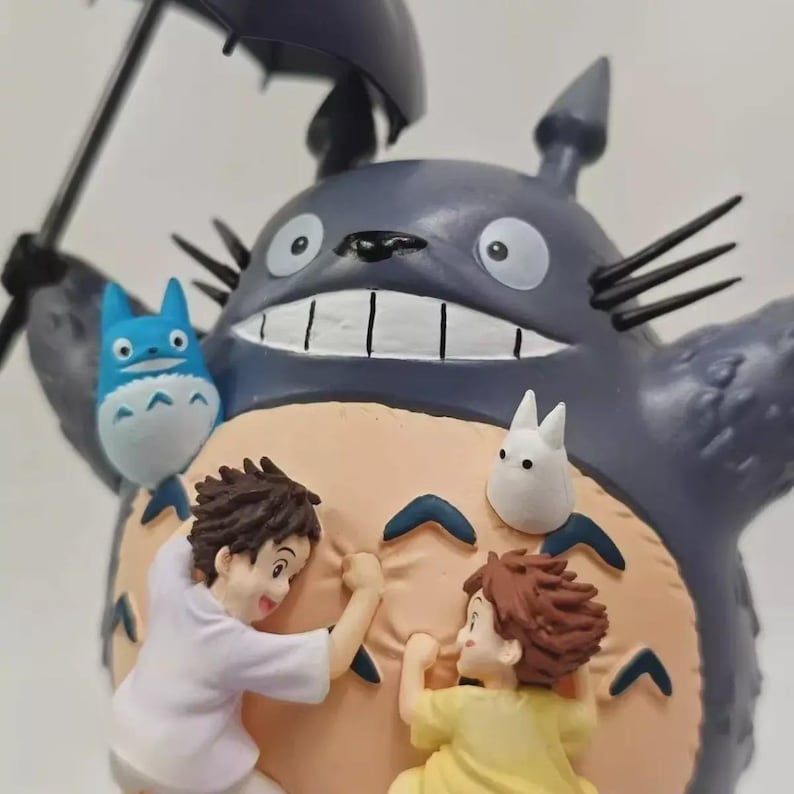 以下が含まれることがあります： アニメ映画「となりのトトロ」のキャラクター、トトロのフィギュア。フィギュアは主にグレーで、お腹はベージュ色で、傘を持っています。2人の子供がキャラクターによじ登っており、近くには2匹の小さな生き物がいます。