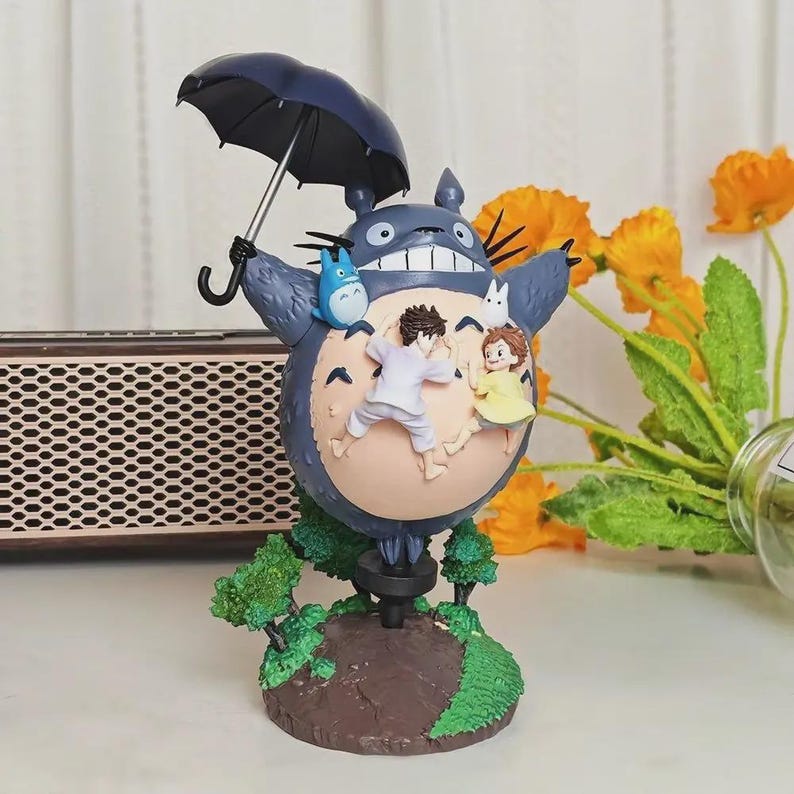 以下が含まれることがあります： 黒い傘を持った大きな青いトトロのキャラクターを特徴とする気まぐれな置物。2つの小さなフィギュアと青い生き物がトトロに取り付けられています。ベースは、緑の木と茶色の土があるミニチュアの風景です。