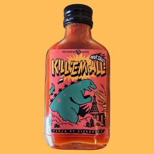 Könnte beinhalten: Eine klare Glasflasche mit scharfer Soße und schwarzem Deckel. Das rosa Etikett zeigt ein grünes Monster, das eine Stadt angreift. Auf dem Etikett steht "KILL'EM ALL" und "HOT SAUCE".