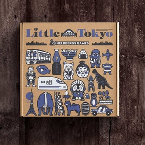 Può includere: Una scatola di cartone marrone con le parole "Little Tokyo" stampate in blu sul davanti. La scatola è piena di varie illustrazioni blu e bianche della cultura giapponese, tra cui un treno, un ristorante di sushi, un mostro simile a Godzilla e una pagoda.
