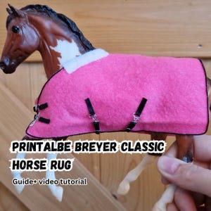Könnte beinhalten: Ein rosa Pferdedecke mit schwarzen Riemen auf einem Spielzeugpferd. Die Decke hat eine weiße Bordüre und ist mit schwarzen Riemen befestigt. Der Text "PRINTALBE BREYER CLASSIC HORSE RUG" ist sichtbar.