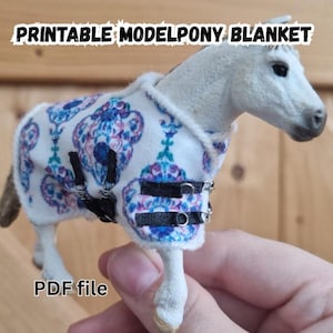 Op de afbeelding: Een wit modelpaard met een patroon deken met blauwe, paarse en turquoise ontwerpen. De deken heeft zwarte bandjes met zilveren gespen. De tekst "PRINTABLE MODELPONY BLANKET" en "PDF file" zijn zichtbaar.