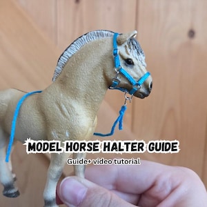 Puede incluir: Un caballo de juguete color canela con crin y cola oscuras que lleva un cabestro azul brillante con anillas metálicas plateadas. El texto MODEL HORSE HALTER GUIDE y Guide+ video tutorial aparece superpuesto en blanco con borde negro.