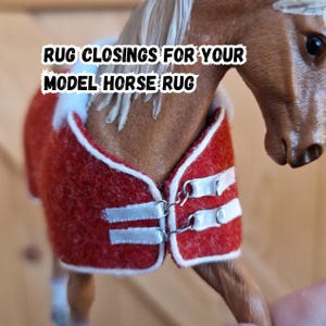 Può includere: Una coperta rossa per cavallo modello con finiture bianche e chiusure argentate. La coperta è su un cavallo modello marrone. Il testo sull'immagine recita "RUG CLOSINGS FOR YOUR MODEL HORSE RUG."
