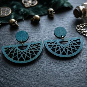 May include: Teal-colored wooden earrings with a half-circle design and intricate cut-out patterns. The earrings have a circular top and hang from gold-colored hooks. The image also includes small skull charms and other decorative elements.