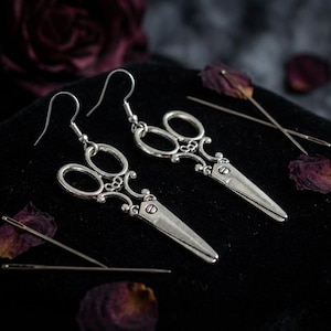 Puede incluir: Pendientes plateados con forma de tijeras y detalles intrincados. Los pendientes se exhiben sobre una superficie negra, acompañados de agujas de coser y pétalos de rosa secos. El diseño presenta una silueta de tijeras clásica con adornos.