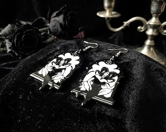 May include: A pair of dangle earrings featuring a black and white illustration of a horned creature with a skull head, set within an arched frame. The earrings have silver-toned metal hooks and a glossy finish, depicting a figure with curved horns.