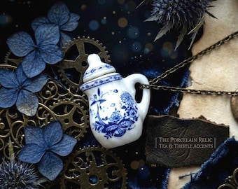 Puede incluir: Un colgante de tetera de porcelana blanca y azul con diseños florales, suspendido de una cadena. El fondo presenta flores azules, engranajes y el texto "The Porcelain Relic Tea & Thistle Accents."