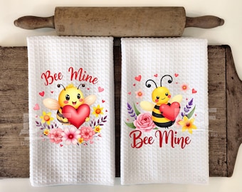 May include: Two white waffle-weave kitchen towels with a Valentine's Day theme. Each towel features a cartoon bee holding a red heart, surrounded by flowers and the text "Bee Mine" in red script. A wooden rolling pin rests above.