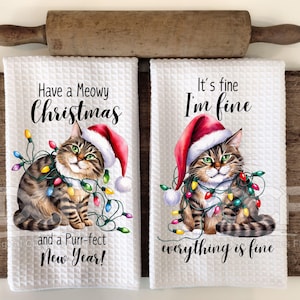 May include: Two white kitchen towels with a cat wearing a Santa hat and Christmas lights. The text on the towels reads "Have a Meowy Christmas and a Purr-fect New Year!" and "It's fine I'm fine everything is fine."