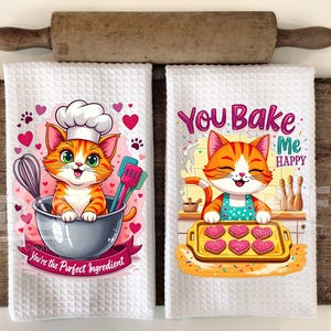 May include: Two white waffle-weave kitchen towels with cartoon cat designs. One features a cat in a chef's hat in a bowl with text "You're the Perfect Ingredient". The other says "You Bake Me HAPPY". A wooden rolling pin is above.