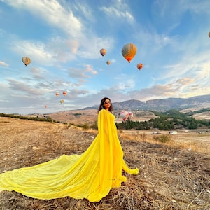 May include: A woman in a bright yellow dress with a long train stands in a field with a view of hot air balloons in the sky.