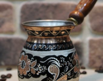 Pode incluir: Uma cafeteira turca tradicional com uma pega de madeira. A cafeteira é feita de cobre e tem um design floral preto e branco com detalhes em cobre.