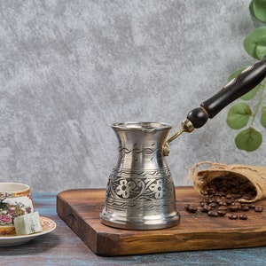 以下が含まれることがあります： 黒い木製のハンドルと花の彫刻が施された、銀色のトルココーヒーポット。 花柄の装飾的なティーカップとソーサー、コーヒー豆の小さなジュート袋、木製のまな板も写っています。