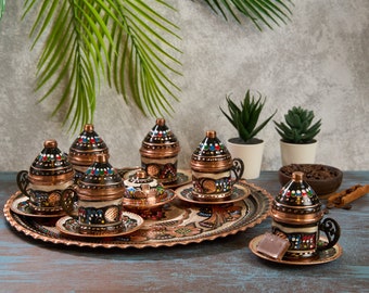 May include: A set of six traditional Turkish coffee cups and saucers with intricate hand-painted designs in black, red, blue, and green. The cups and saucers are made of copper and have a shiny finish. The set is displayed on a copper tray with a decorative border.