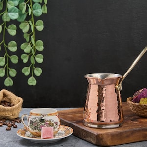 May include: A copper Turkish coffee pot with a long handle sits on a wooden cutting board. A small white cup with a floral design and a pink Turkish delight sits on a white saucer. A burlap bag filled with coffee beans sits on a gray surface. A small woven basket filled with colorful Turkish delights sits on the cutting board.