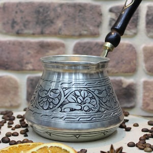 May include: A silver Turkish coffee pot with an ornate floral design and a black wooden handle. The pot is sitting on a wooden surface with coffee beans, star anise, and dried orange slices.