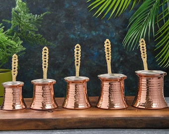 Pode incluir: Um conjunto de cinco cafeteiras cezve de cobre com bordas prateadas e alças douradas ornamentadas. As cafeteiras de cobre martelado são exibidas em uma tábua de madeira.