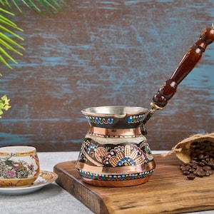 Puede incluir: Una cafetera turca tradicional con cuerpo de cobre y mango de madera. La cafetera está decorada con coloridos patrones florales y está sobre una tabla de cortar de madera. Hay una taza pequeña blanca y un platillo con un diseño floral al lado de la cafetera.