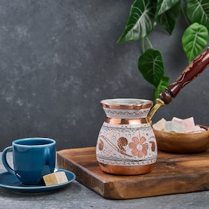 May include: A copper and white Turkish coffee pot with a wooden handle, a blue coffee cup and saucer, and a bowl of Turkish delight on a wooden cutting board.