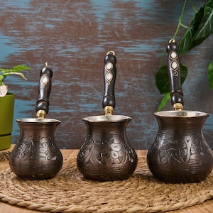 May include: Three traditional Turkish coffee pots with ornate floral designs and wooden handles. The pots are made of copper and have a dark brown finish.