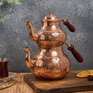 Könnte beinhalten: Ein traditionelles türkisches Teeset mit zwei Kupferkesseln, die übereinander gestapelt sind. Die Kessel haben aufwendige Blumendekore und Holzgriffe. Das Set steht auf einer Holzoberfläche mit einem Glas Tee und getrockneten Orangenscheiben.