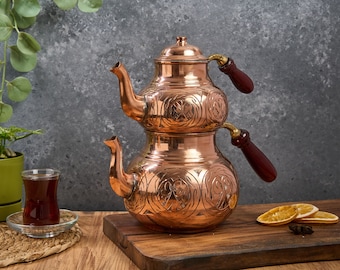 May include: A traditional Turkish tea set with two copper kettles, one stacked on top of the other. The kettles have intricate floral designs and wooden handles. The set is on a wooden surface with a glass of tea and dried orange slices.