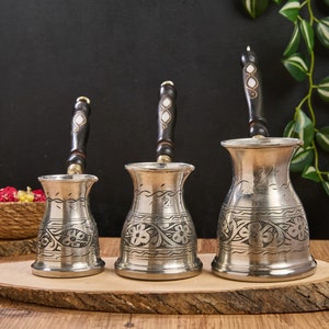 May include: Three silver Turkish coffee pots with black wooden handles and intricate floral designs. The pots are arranged on a wooden surface.