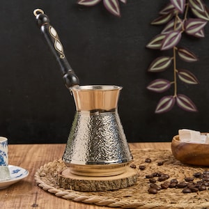 May include: A silver Turkish coffee pot with a hammered finish and a black wooden handle with white accents. The pot is sitting on a woven placemat with coffee beans scattered around it. A bowl of Turkish delight is in the background.