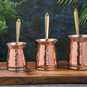 May include: Three hammered copper coffee pots with brass handles on a wooden surface. The pots are different sizes.