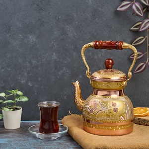 May include: A gold and copper teapot with intricate floral designs and a wooden handle. A glass of tea sits on a wooden table next to the teapot.