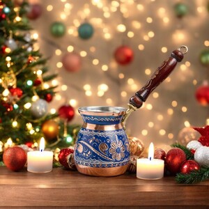 May include: A traditional Turkish coffee pot with a blue and copper design and a wooden handle. The pot is sitting on a wooden table with candles and Christmas ornaments.