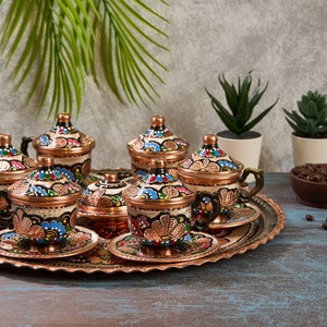 May include: A set of six Turkish coffee cups and saucers featuring intricate hand-painted floral designs in shades of blue, red, and green. The cups and saucers are made of copper and have a shiny finish. The set is arranged on a copper tray with a scalloped edge.