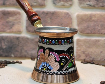 Pode incluir: Uma cafeteira turca tradicional com uma pega de madeira e um corpo de cobre. A cafeteira é decorada com padrões florais coloridos e está sobre uma superfície de madeira com grãos de café, anis estrelado e fatias de laranja.