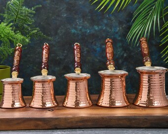 Pode incluir: Conjunto de cinco bules de café de cobre com alças de madeira. Os bules têm um acabamento martelado e estão dispostos numa superfície de madeira.