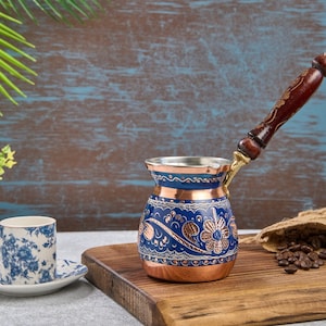 Könnte beinhalten: Eine türkische Kaffeekanne aus Kupfer und Blau mit Holzgriff, floralem Design und passender Tasse und Untertasse. Kaffeebohnen quellen aus einem Jutesack auf einem Holzbrett.