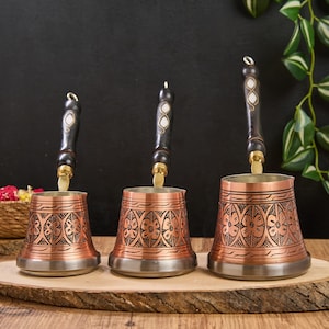 Puede incluir: Tres cafeteras de cobre con diseños florales y mangos de madera oscura. Las jarras se exhiben sobre una tabla de madera, mostrando su acabado de cobre pulido y detalles intrincados, para café turco.