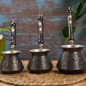 Könnte beinhalten: Drei traditionelle türkische Kaffeekannen mit verzierten Gravuren und Holzgriffen. Die Kannen sind aus Kupfer und haben eine dunkelbraune Oberfläche.