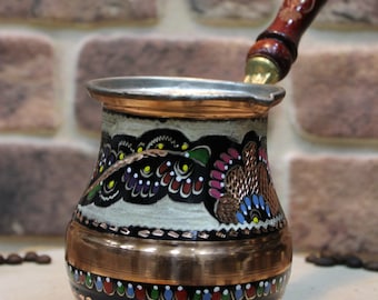 Pode incluir: Uma cafeteira turca de cobre com uma pega de madeira. A cafeteira é decorada com padrões florais coloridos e uma borda preta.