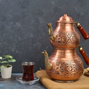 May include: A traditional Turkish tea set with a copper teapot and two cups. The teapot has intricate floral designs and is stacked on top of a larger copper pot. The cups are clear glass and are sitting on a saucer. The tea set is on a rustic brown surface.