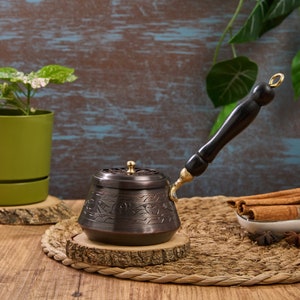 Puede incluir: Una cafetera turca tradicional con un mango de madera negro y una intrincada metalurgia. La cafetera está sentada sobre una base de madera con una estera de paja tejida debajo. Hay palitos de canela y anís estrellado en la estera.